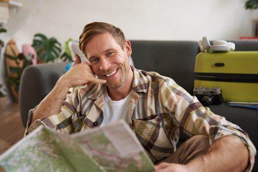 Smiling young man with travel map, shows mobile phone call me gesture, sits with luggage in living room, goes on holiday.