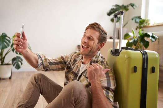 Cheerful smiling man with suitcase, video chats using smartphone, takes selfie, goes on holiday.
