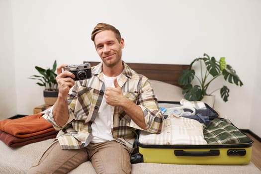 Portrait of handsome man going on vacation, packing suitcase, holding camera, travel photographer going on holiday.