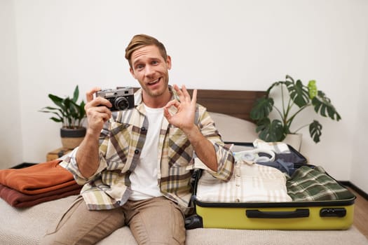 Portrait of happy young man in hotel, unpacking his suitcase on vacation, holding camera, showing okay, good hand sign, approve something, recommending holiday destination.