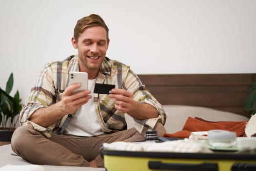 Portrait of young man booking tickets, going on holiday, holding credit card and smartphone, sitting with mobile phone near suitcase filled with clothes, ready for vacation trip.