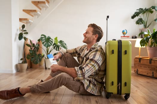 Portrait of handsome young man, tourist sitting with his suitcase, holding passport and flight tickets, going on holiday.