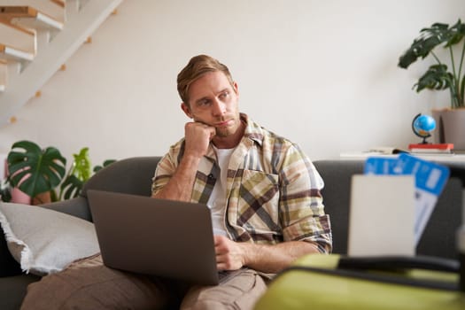 Portrait of upset young man, boyfriend plans holiday trip, has two plane tickets abroad for travelling on holiday, works on laptop.