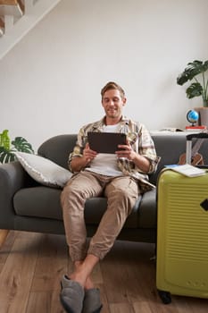 Vertical shot of man, tourist with suitcase, looking at digital tablet, reading about travel destination, booking hotel on gadget. Tourism concept