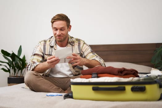 Portrait of handsome man, tourist, looking at his holiday tickets, booked vacation with travel agency, sitting on bed and packing clothes in suitcase.