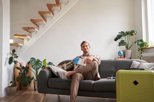 Portrait of handsome young man, bought tickets for plane, going on holiday, sitting on sofa with packed suitcase, waiting for taxi to airport. Copy space