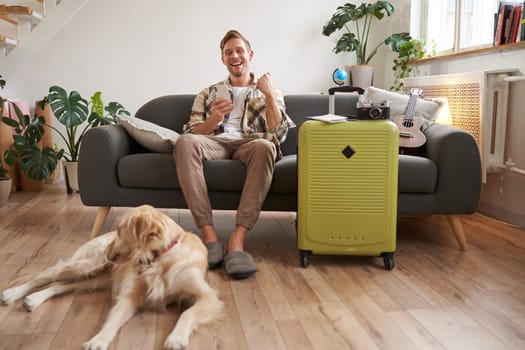 Image of excited man, tourist with a dog, sits in living room with packed suitcase and travel camera, guy saying yes and celebrating, pleased with prices online, booking pet-friendly hotel.