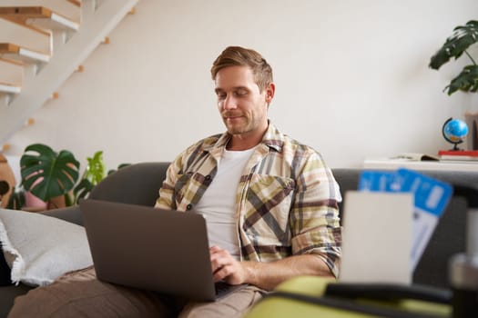 Portrait of handsome young man, has two tickets and passport, goes online on laptop, booking hotel and planning his holiday, sitting in living room.