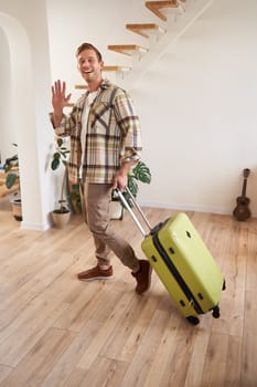 Full length photo of happy young man, tourist going on holiday, traveller leaving apartment with suitcase, waving hand at camera to say goodbye. Copy space