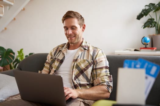 Portrait of young handsome man sitting with laptop, has passport and two plane tickets prepared, planning his journey on computer, booking hotels and restaurants online for the trip.