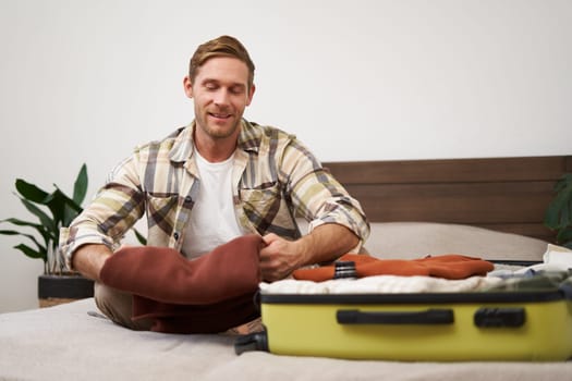 Portrait of handsome young man, tourist unpacking clothes in hotel room, staying in hostel on vacation, travelling, sitting with luggage on bed.