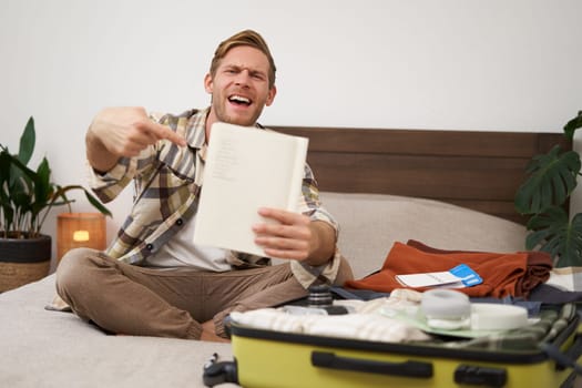 Travelling and tourism. Young man sits on bed, shows his list of items, demonstrates notebook, packs clothes, prepares to go on holiday, goes on vacation, assembles luggage.