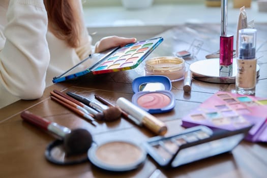Close-up of cosmetic products, brushes, mirror on table, hands of young woman with eyeshadow palette. Cosmetics, beauty, makeup concept