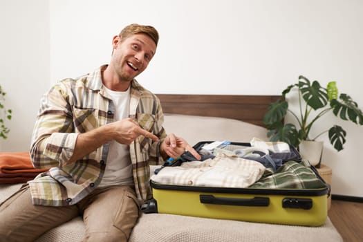 Handsome smiling man, pointing at his suitcase with clothes, invites for holiday, encourages to pack and go on vacation, sits on bed with luggage.