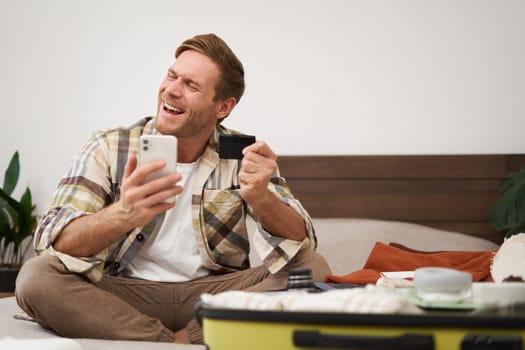 Cheerful tourist, guy with credit card and mobile phone, paying for holiday tickets, sitting on bed with suitcase, packing and going on holiday.