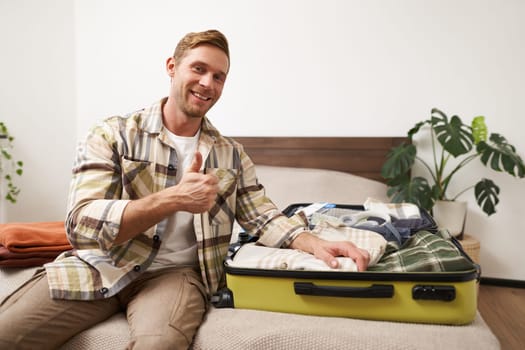 Portrait of handsome smiling man, tourist shows thumbs up, sits with opened suitcase, packs his clothes inside luggage and looks pleased, goes on vacation, prepares for a holiday.