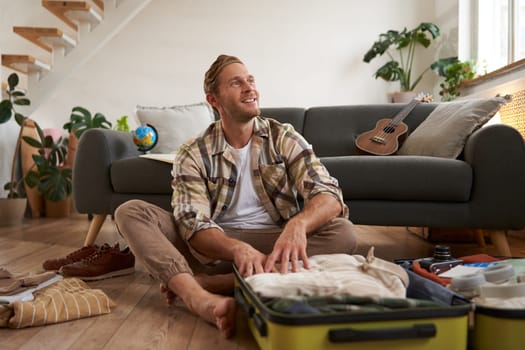 Happy young traveller, man sits with luggage on floor, looking aside and smiling, thinking about his future holiday, going on vacation, packing suitcase.