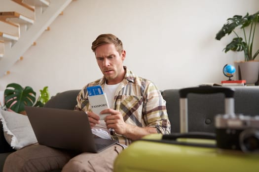 Portrait of grumpy man, holding tickets and passport, has laptop opened, looking concerned and angry, sitting with suitcase on sofa.