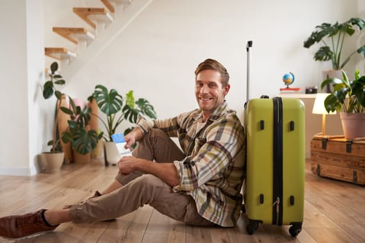 Portrait of smiling handsome man, sitting with suitcase on floor, holding passport and flight tickets, going on tour vacation.