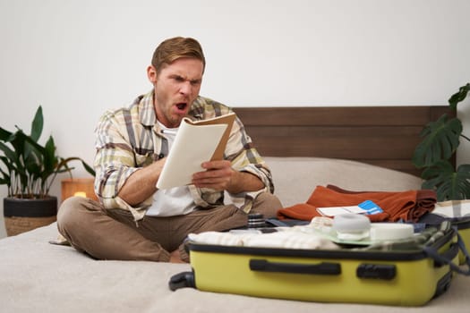 Portrait of angry man, packing suitcase, looking frustrated at notebook with check list of items for holiday, preparing to go on vacation. Tourist and people concept