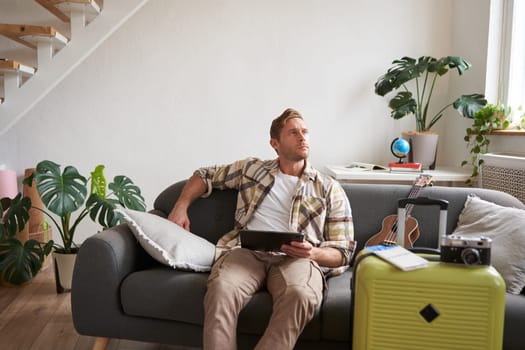 Image of young man with digital tablet, tourist sitting on sofa with suitcase packed and ukulele, thinking, looking aside thoughtful, planning holiday trip. Tourism concept