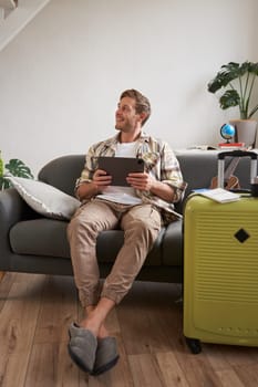 Vertical shot of handsome man in living room, sitting with suitcase and digital tablet, looking aside and smiling, booking hotel for holiday trip, going on vacation, planning the travel.