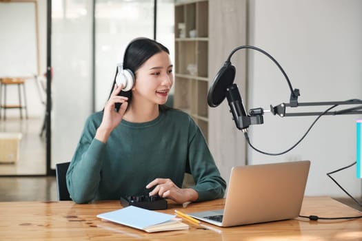 A woman is sitting at a desk with a laptop and a microphone. She is wearing headphones and she is recording a podcast or a voiceover. The room has a modern and professional atmosphere