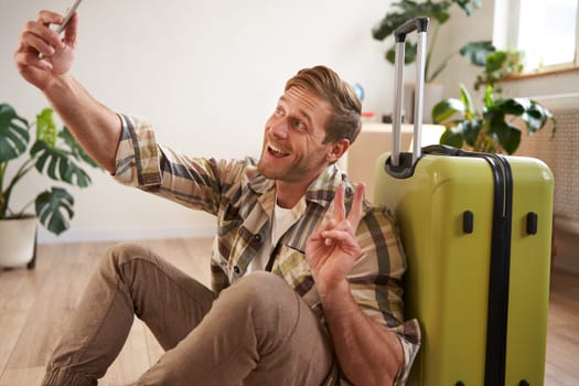 Portrait of smiling handsome man with suitcase, taking selfie with luggage, posting on social media about going on holiday. Tourism and blogging concept