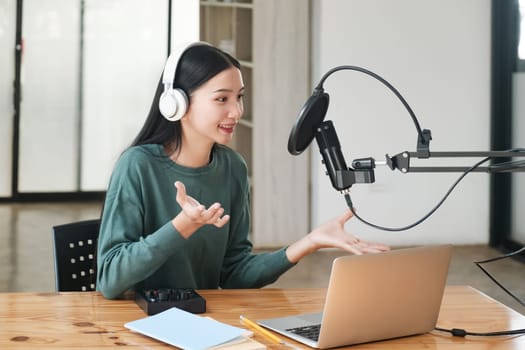 A woman is sitting at a desk with a laptop and a microphone. She is wearing headphones and she is talking into the microphone. The scene suggests that she is recording a podcast or a voiceover session