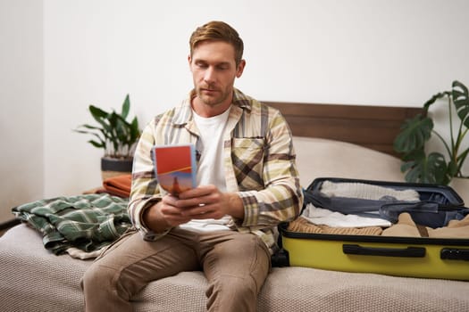 Portrait of young man, tourist is packing his luggage, looking at tour guide, travel map, sitting on bed with his clothes in a suitcase, Travelling and tourism concept