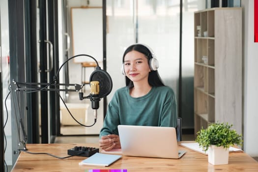 A woman is sitting at a desk with a microphone and a laptop. She is wearing headphones and smiling. The scene suggests that she is recording a podcast or a voiceover session