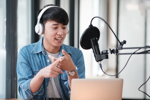 A man wearing headphones and a blue shirt is talking into a microphone. He is smiling and pointing to something on a laptop. The scene suggests that he is giving a presentation or giving instructions
