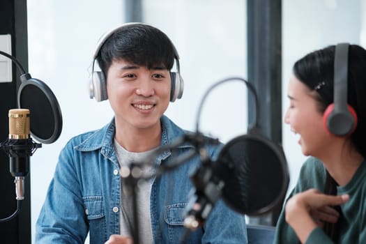 Two people are sitting in a studio with microphones and headphones on. One of them is smiling and the other is laughing. Scene is lighthearted and fun