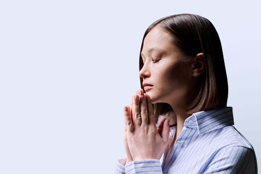 Close-up profile view of young woman with folded hands on white background, copy space. Serious female with closed eyes worried praying thinking. Mental health psychology psychotherapy feelings stress