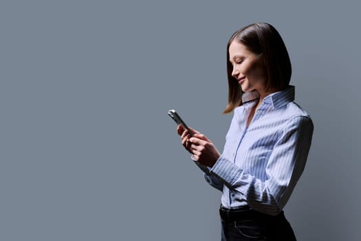 Young happy woman with smartphone in hands on grey background profile view copy space. Smiling female looking at phone screen texting reading. Using mobile applications for work business study leisure