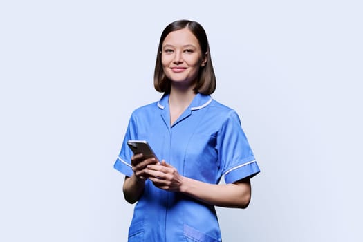 Smiling young female nurse using smartphone, looking at camera on white studio background. Mobile apps applications, technologies in medical services, health, professional assistance, medical care