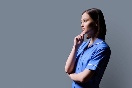 Portrait of young confident serious female nurse looking in profile on gray studio background, copy space. Medical services, health, professional assistance, medical care concept