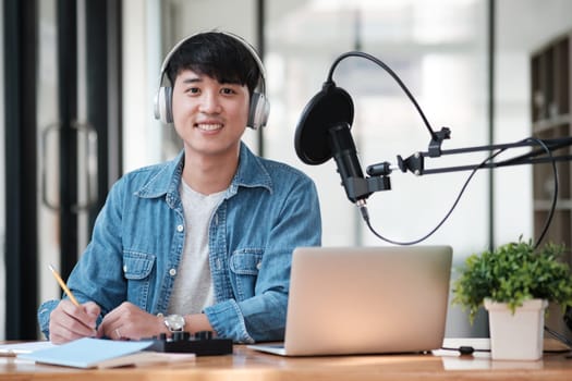 A man wearing headphones and a blue shirt is sitting at a desk with a laptop and a microphone. He is smiling and he is happy