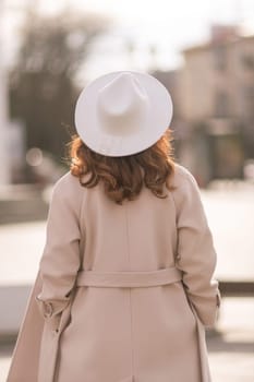 Back view of happy woman wearing hat and coat walking down street on sunny spring day. People, lifestyle, travel and vacations concept.