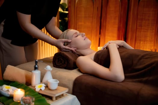 Caucasian woman enjoying relaxing anti-stress head massage and pampering facial beauty skin recreation leisure in warm candle lighting ambient salon spa in luxury resort or hotel. Quiescent