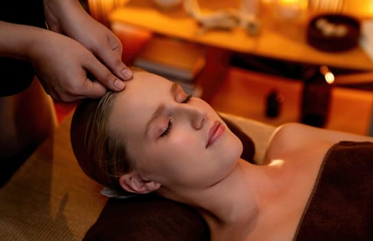 Caucasian woman enjoying relaxing anti-stress head massage and pampering facial beauty skin recreation leisure in warm candle lighting ambient salon spa in luxury resort or hotel. Quiescent