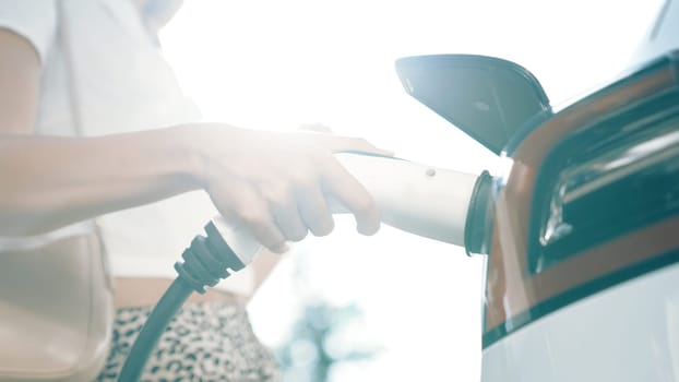 Sunlight backlit brighten young woman travel with EV electric car charging in green sustainable city outdoor garden in summer. Green urban sustainability lifestyle by electric vehicle innards