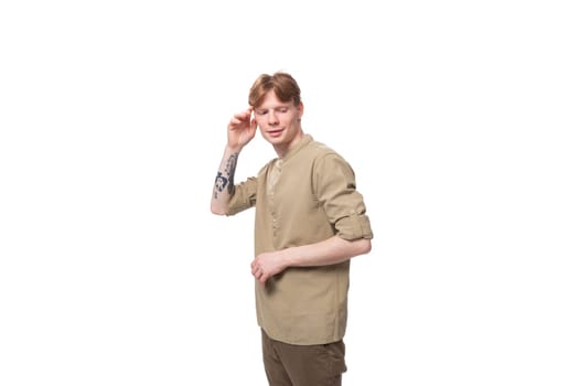 young well-groomed caucasian man with red hair is dressed in a khaki shirt and brown trousers on a white background.