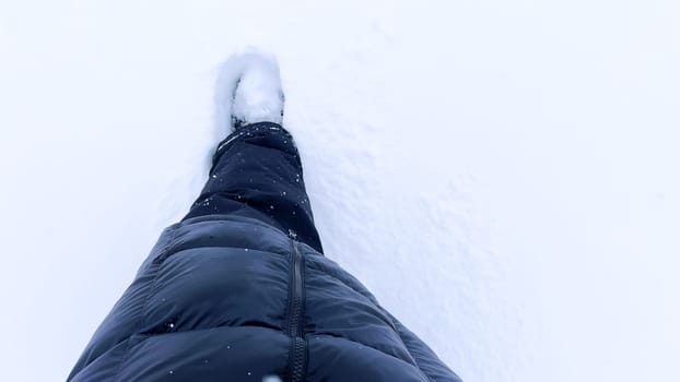 The view focuses on the lower half of a person, stepping into a vast expanse of pristine snow, leaving a single trail behind. The simplicity of the winter attire contrasts starkly with the endless white canvas below.