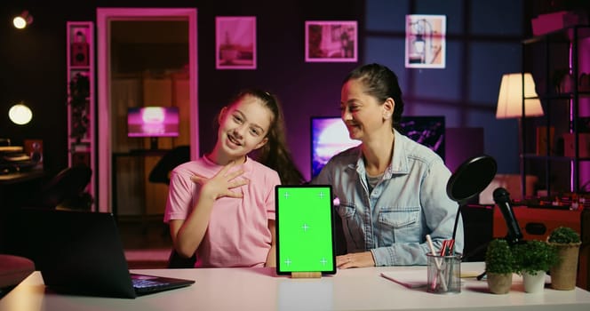 Cute kid filming internet show helped by her mother, promoting green screen tablet from sponsoring brand. Little girl and her mom do influencer marketing, urging followers to purchase mockup device