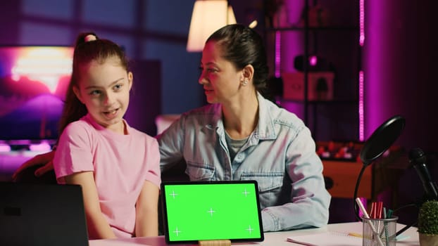 Cheerful child and loving mom record chroma key tablet review on online platforms. Young smiling daughter and parent presenting isolated screen device, filming in home studio with pink neon lighting