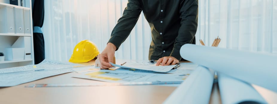 A portrait of professional male engineer shows mistake point in blueprint on messy table with architectural plan and yellow helmet placed at modern office. Closeup. Focus on hand. Delineation.