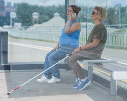 An elderly blind woman and a pregnant woman are sitting at a bus stop and waiting for the bus