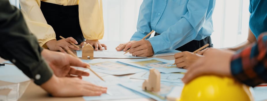 Professional architect team and engineer discuss about architectural project on meeting table with safety helmet, wooden block and architectural document scatter around. Closeup. Delineation.