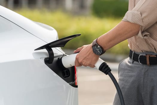 Young man put EV charger to recharge electric car's battery from charging station in city commercial parking lot. Rechargeable EV car for sustainable environmental friendly urban travel. Expedient